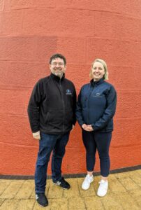 Two people standing together, in front of an orange wall. 
