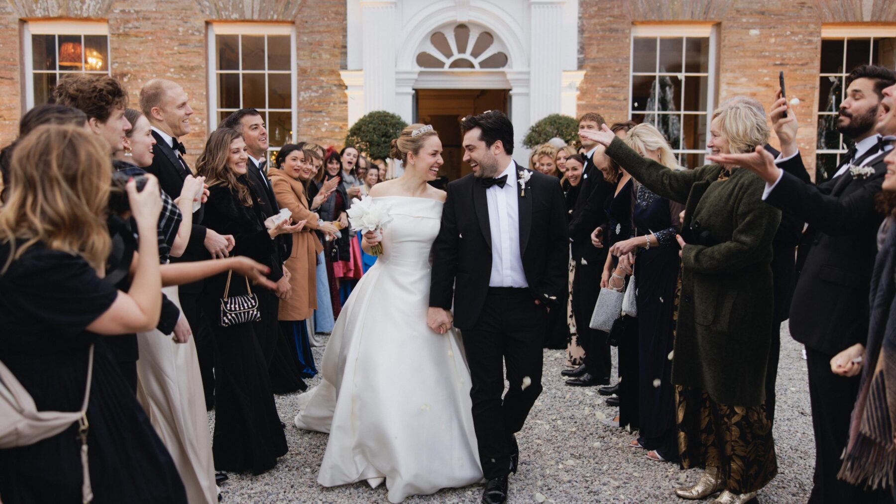 A newly married couple walking between two rows of celebrating guests, looking at each other as confetti is thrown around them.