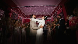 A newly married couple sharing their first dance on the dancefloor, embracing under a structured fairy light canopy while surrounded by guests filming and celebrating.