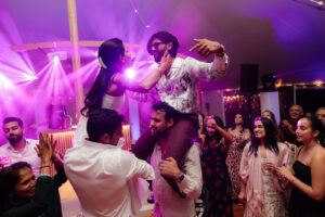A bride celebrating their wedding on someone’s shoulders facing another guest on shoulders, under purple lighting with a packed wedding dance floor around them.